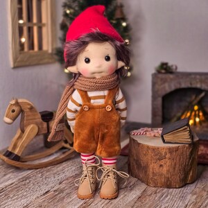 May include: A handmade doll dressed in a red hat, brown scarf, and brown overalls. The doll has a striped shirt, freckles, and tan boots. A wooden rocking horse and a small fireplace are in the background.