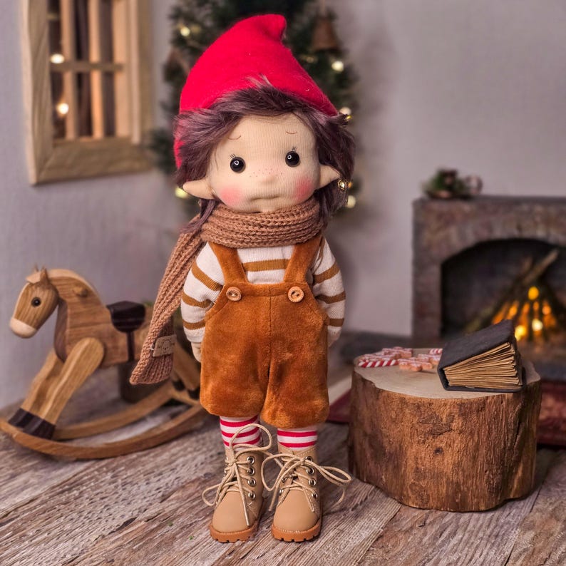 May include: A handmade doll dressed in a red hat, brown scarf, and brown overalls. The doll has a striped shirt, freckles, and tan boots. A wooden rocking horse and a small fireplace are in the background.