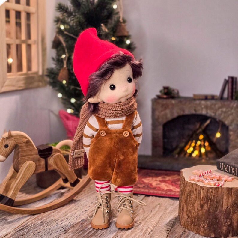 May include: A handmade doll dressed in a red hat, brown overalls, and a striped shirt. The doll has a brown scarf and beige boots. The doll is standing in front of a Christmas tree and a wooden rocking horse.