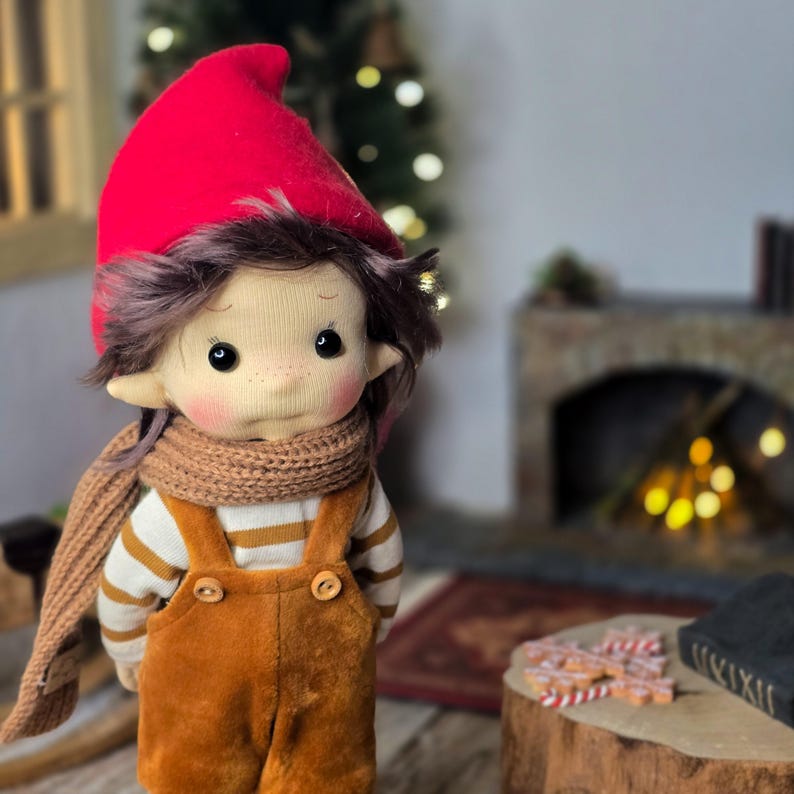 May include: A handmade doll dressed in a red pointed hat, brown scarf, and orange overalls. The doll has a white and tan striped shirt, brown hair, and rosy cheeks. The doll is standing in front of a fireplace and Christmas tree.