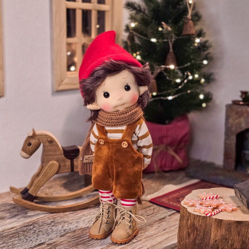 May include: A handmade doll dressed in a red hat, brown overalls, and a striped shirt. The doll has a brown scarf and boots. A wooden rocking horse and a small Christmas tree are in the background.
