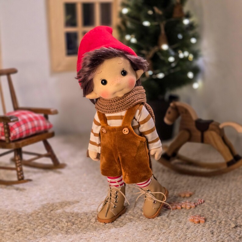 May include: A handmade doll dressed in a red hat, brown hair, and a tan and white striped shirt. The doll wears brown overalls, a brown scarf, and tan boots. The doll has rosy cheeks and freckles. A Christmas tree and rocking horse are in the background.