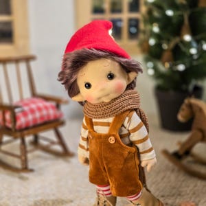May include: A handmade doll with a red pointed hat, brown hair, and rosy cheeks. The doll wears a striped shirt, brown overalls, a knitted scarf, and boots. The doll is standing in front of a Christmas tree and a rocking horse.