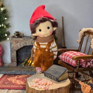May include: A handmade doll dressed in a red hat, brown overalls, and a striped shirt. The doll has a scarf and is standing in front of a fireplace. A wooden stump holds gingerbread cookies and a book. A rocking chair with a plaid pillow is in the background.