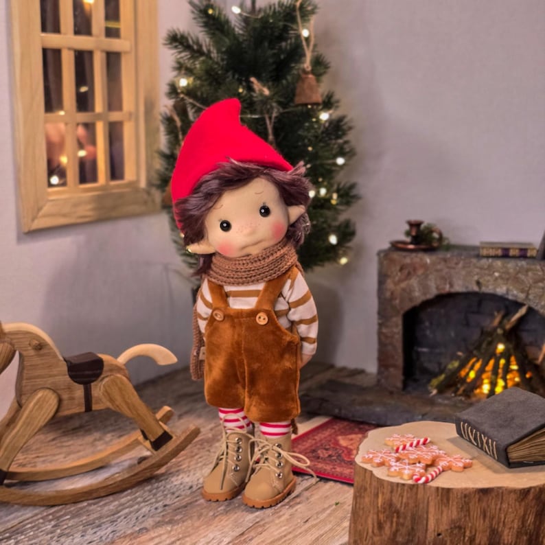May include: A handmade doll dressed in a red hat, brown overalls, and a striped shirt. The doll has a scarf, boots, and rosy cheeks. The doll is standing in a room with a Christmas tree and a wooden rocking horse.