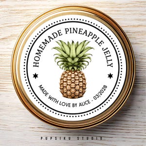以下が含まれることがあります： 黒文字と白黒のパイナップルのイラストが描かれた白い丸いラベル。ラベルには「自家製パイナップルゼリー」と「Made with Love by Alice - 03/2028」と書かれています。