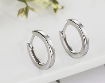 May include: A pair of small, polished silver-toned hoop earrings with a hinged closure mechanism. The earrings feature a smooth, rounded surface and a reflective metallic finish, presented as a matching set of circular jewelry pieces.