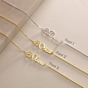 May include: Three gold necklaces with a paw print charm and a personalized name. The names are Daisy, Oscar, and Olivia. The necklaces are shown in three different fonts.