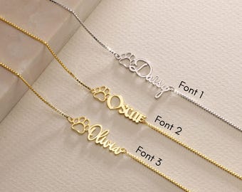 Può includere: Tre collane d'oro con un ciondolo a forma di zampa e un nome personalizzato. I nomi sono Daisy, Oscar e Olivia. Le collane sono mostrate in tre font diversi.