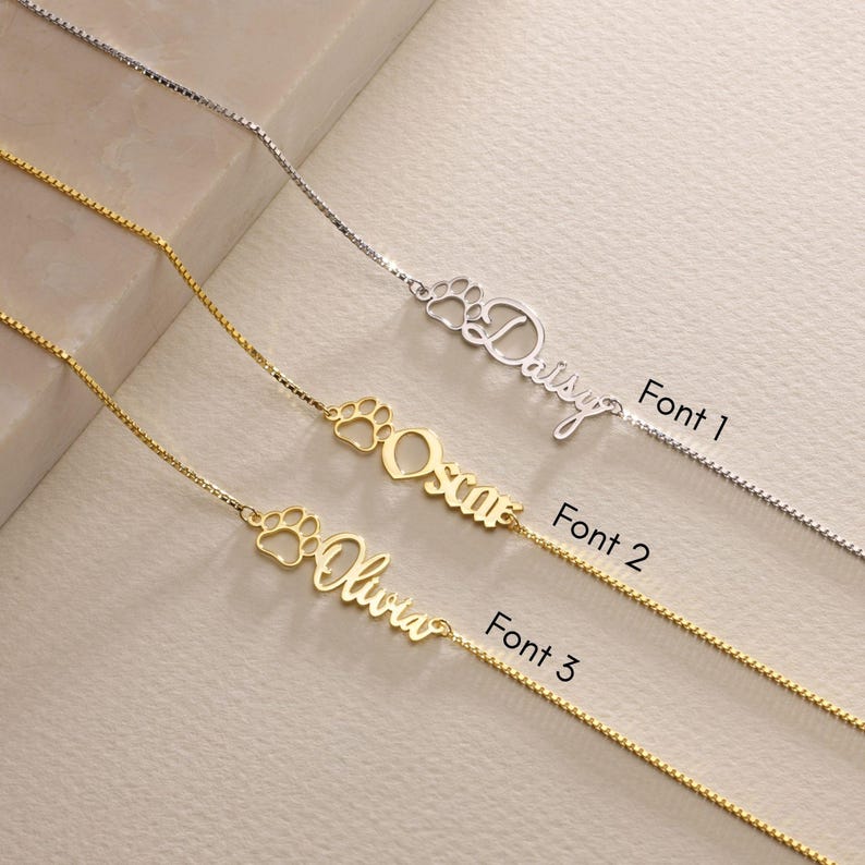 May include: Three gold necklaces with a paw print charm and a personalized name. The names are Daisy, Oscar, and Olivia. The necklaces are shown in three different fonts.