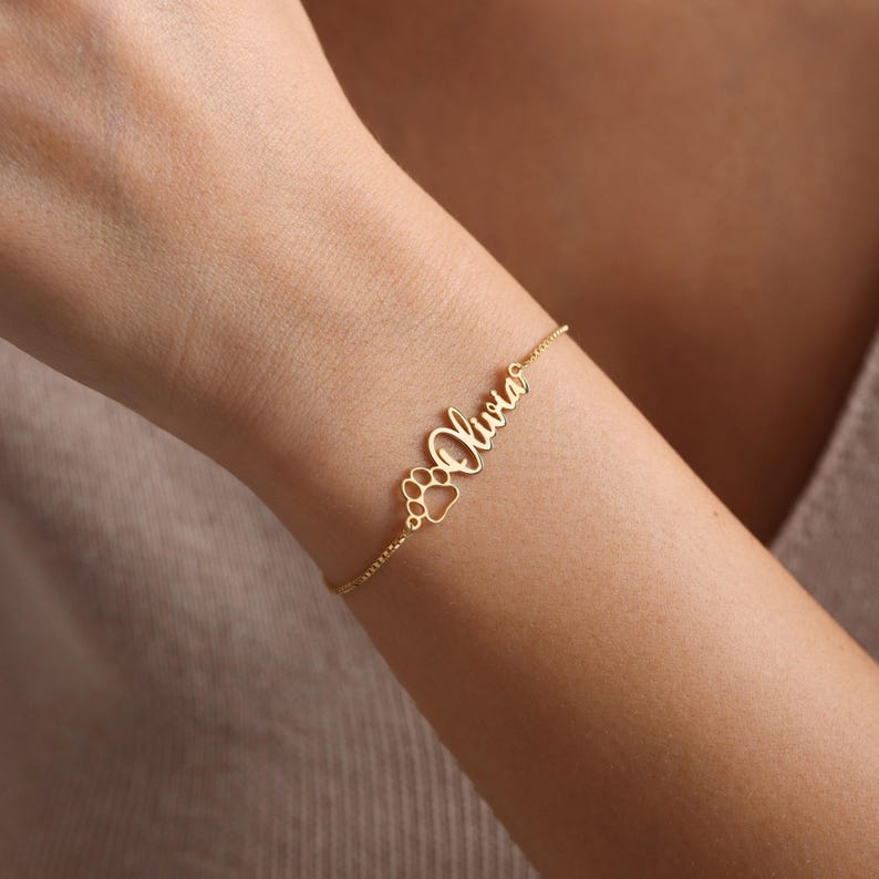 May include: Gold bracelet with the name "Olivia" and a paw print charm.