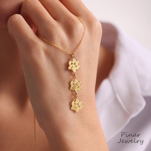 May include: Gold necklace with three paw print charms, each engraved with a letter. The necklace is on a hand wearing a white shirt.