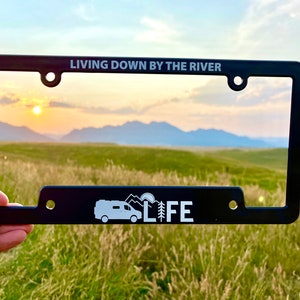 May include: Black metal license plate frame with the text "LIVING DOWN BY THE RIVER" on the top and a white silhouette of a camper van with the text "LIFE" on the bottom. The frame is held in front of a scenic landscape with mountains and a sunset.