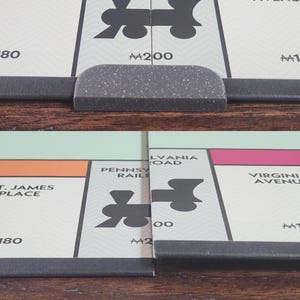 May include: Close-up of a Monopoly board game. The board shows property cards with street names like St. James Place and Virginia Avenue. The game pieces are visible, along with the board's edge.