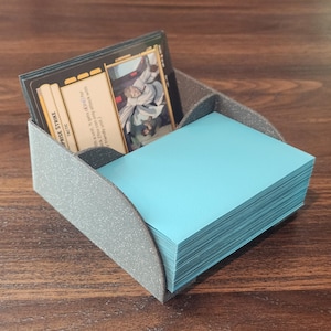 May include: A gray card holder with a stack of light blue cards and a few game cards. The game cards have images and text. The holder is on a dark wood surface.