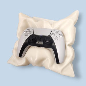 May include: A white and black PlayStation 5 DualSense wireless controller rests on a cream-colored, pillow-like form against a light blue background. The controller features a central touchpad, directional buttons, and analog sticks. The overall aesthetic is modern and sleek.