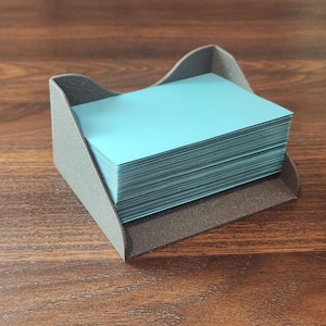 May include: A stack of teal-colored cards sits in a gray and brown card holder. The holder has a curved design on the sides and is made of a textured material. The cards are neatly stacked, suggesting organization and a professional setting.