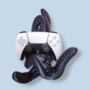 May include: A white PlayStation 5 controller is held by a dark blue, tentacled octopus-like sculpture. The controller has black joysticks and button symbols. The sculpture has a textured surface and is set against a light blue background.
