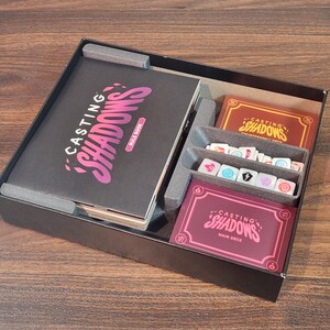 May include: A board game box with the title "Casting Shadows" on the rule book and card decks. The box contains a rule book, card decks, dice, and other game components. The color scheme includes black, burgundy, and pink.