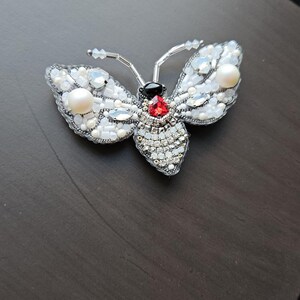 Peut inclure: Une broche en forme de papillon en perles argentées et blanches avec une gemme rouge et noire au centre.