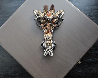 Peut inclure: Broche girafe artisanale, ornée de perles marron, dorées et noires. La girafe a un visage détaillé avec de petites lunettes et un nœud papillon. La broche est posée sur une surface grise.