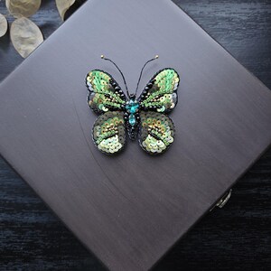 Puede incluir: Un broche de mariposa verde y negro con lentejuelas y pedrería. El broche está sobre una superficie marrón oscura.