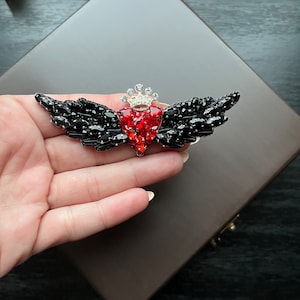 Puede incluir: Un corazón rojo adornado con joyas con una corona plateada, rodeado de alas negras, hecho de diamantes de imitación. El corazón es un broche.