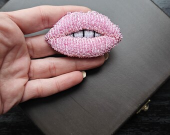 May include: A pink beaded brooch shaped like a pair of lips. The lips are closed and have a white beaded line across the center.