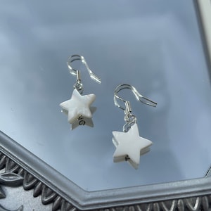 Pode incluir: Um par de brincos de estrela prateados com detalhes de pérola branca. Os brincos estão pendurados em uma superfície de espelho.