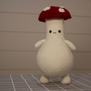 May include: A handmade crochet mushroom figure with a red cap and white spots. The mushroom has a cream-colored body, arms, and legs, and a small, smiling face. The whimsical design is perfect for home decor.