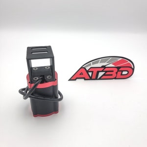 Op de afbeelding: Een zwart-rode 3D-geprinte motor met een zwarte draad eraan bevestigd. De motor heeft een zwart frame en een rode cilindrische behuizing. De motor staat op een wit oppervlak. Het logo "AT3D" is zichtbaar op de achtergrond.
