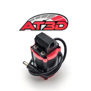 May include: A black and red electronic component with a black bracket secured by screws. A black cable with a DC connector is attached. The logo "AT3D" in red and white is in the background.