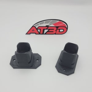 May include: Two black 3D printed wall mounts with a square base and a rectangular opening. The mounts are designed to hold a device or object.