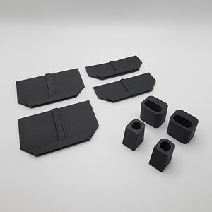 以下が含まれることがあります： 黒色のプラスチック製の3Dプリントパーツのセット。中央に盛り上がった隆起がある平らなパーツが4つと、上部に穴が開いた小さなブロックが4つ含まれています。