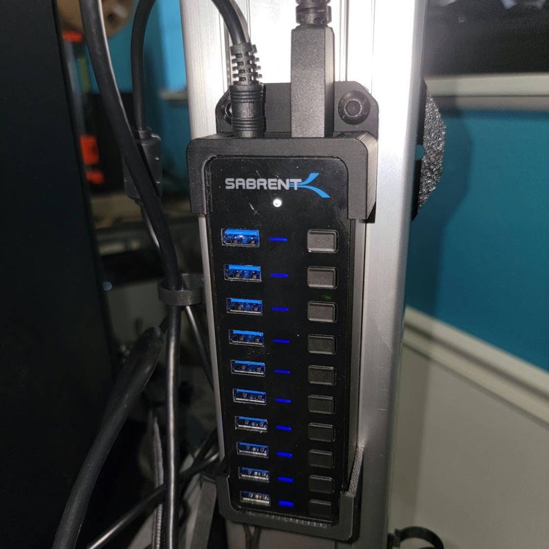 May include: A black USB hub with 7 ports and a blue LED light. The hub is mounted on a silver metal surface. The brand name "SABRENT" is printed on the front of the hub.