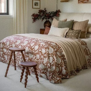 Può includere: Una camera da letto con un letto rivestito con un copripiumino a motivi floreali bianchi e marroni e una coperta beige. Due sgabelli in legno con un design attorcigliato sono in primo piano. Il letto ha una testiera bianca ed è decorato con cuscini in varie tonalità di marrone e verde.