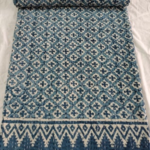 May include: A rolled-up, blue and white patterned quilt. The quilt has a repeating floral design and a decorative border. The stitching is visible, adding texture to the fabric. The quilt is made of soft material and is suitable for a bed or sofa.