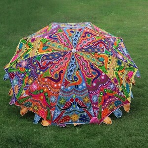 Puede incluir: Un paraguas pintado a mano con colores vibrantes y un dise&ntilde;o intrincado. El paraguas presenta una variedad de colores, incluyendo rojo, azul, amarillo y verde, con un patr&oacute;n de p&aacute;jaros y motivos florales. El paraguas est&aacute; abierto y sobre hierba verde.
