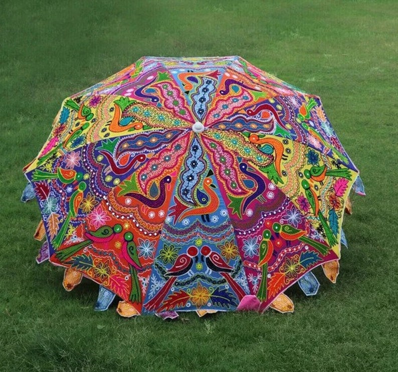 Puede incluir: Un paraguas pintado a mano con colores vibrantes y un dise&ntilde;o intrincado. El paraguas presenta una variedad de colores, incluyendo rojo, azul, amarillo y verde, con un patr&oacute;n de p&aacute;jaros y motivos florales. El paraguas est&aacute; abierto y sobre hierba verde.