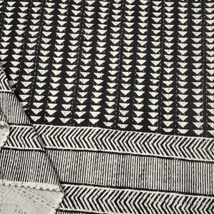 Puede incluir: Colcha estampada en blanco y negro con un diseño geométrico. La colcha presenta filas de triángulos blancos sobre un fondo negro, con un borde de rayas verticales y en zigzag. Los bordes están cosidos con hilo blanco.