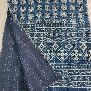 Könnte beinhalten: Eine gefaltete Steppdecke mit indigo-blauen und weißen Mustern. Die Oberseite zeigt ein sich wiederholendes kreisförmiges Design mit geometrischen und linearen Mustern darunter. Die Rückseite ist einfarbiges Indigo-Blau mit weißen Nähten. Die Steppdecke ist aus weichem Stoff.
