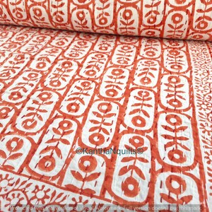 May include: A rolled-up, white and orange floral patterned quilt. The quilt features a repeating design of stylized flowers and leaves, with a border of floral and striped patterns. The fabric is a soft, textured material.