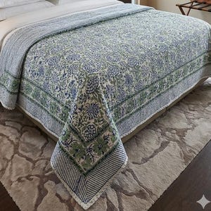 May include: A floral patterned quilt in shades of blue and green, draped over a bed. The quilt features a repeating floral design on a cream background, with striped borders. The bed is made with white sheets and a neutral-colored rug is beneath.