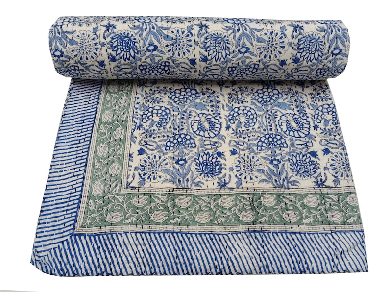 May include: A rolled-up, floral-patterned quilt with a cream background and blue floral designs. The quilt has a blue and green border with a striped edge. The design features a repeating pattern of flowers and leaves.