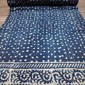 May include: A blue and white kantha quilt with a white polka dot pattern and a floral border. The quilt is folded and rolled up.