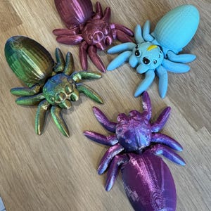 May include: Four 3D printed spiders in different colours: purple, blue, green, and red. The spiders have a shiny, iridescent finish.
