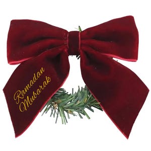 May include: A deep red velvet bow with gold script reading "Ramadan Mubarak". The bow is set on a small green wreath, a decorative item for the occasion.