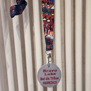 Op de afbeelding: Een blauw koord met Spider-Man-afbeeldingen, een zilverkleurige metalen sluiting en een ronde tag. De tag leest "Bravo Luka, Roi du Trône 04/08/2025" in rode tekst.