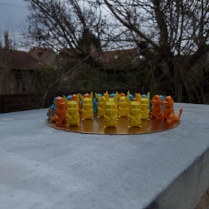 May include: A group of 15 small, origami-style figures arranged in a circle on a gold surface. The figures are yellow, orange, and blue, and they appear to be characters from the Pokemon franchise.