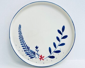 Può includere: Un piatto in ceramica bianca con un design floreale blu e rosso. Il piatto ha un bordo blu e un centro bianco con disegni floreali blu e rossi.